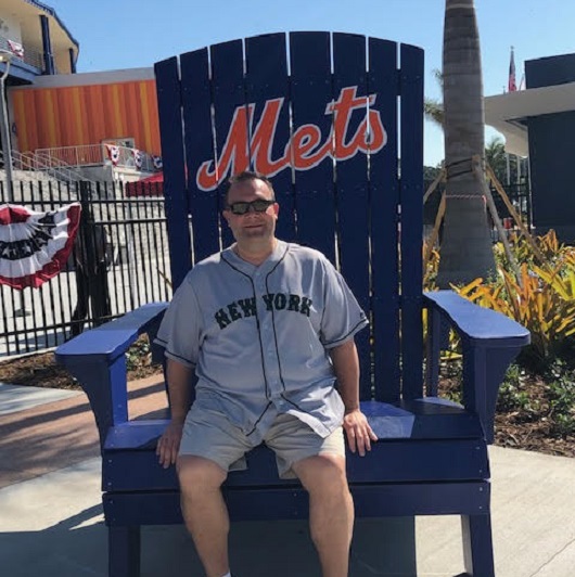 Patient, Jim Bleemer, wearing a Mets jersey, sitting in a Mets branded chair.