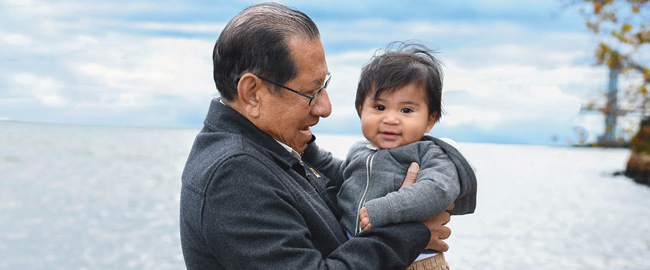 Elderly man with glasses and a dark gray jacket holding a baby in a gray hoodie