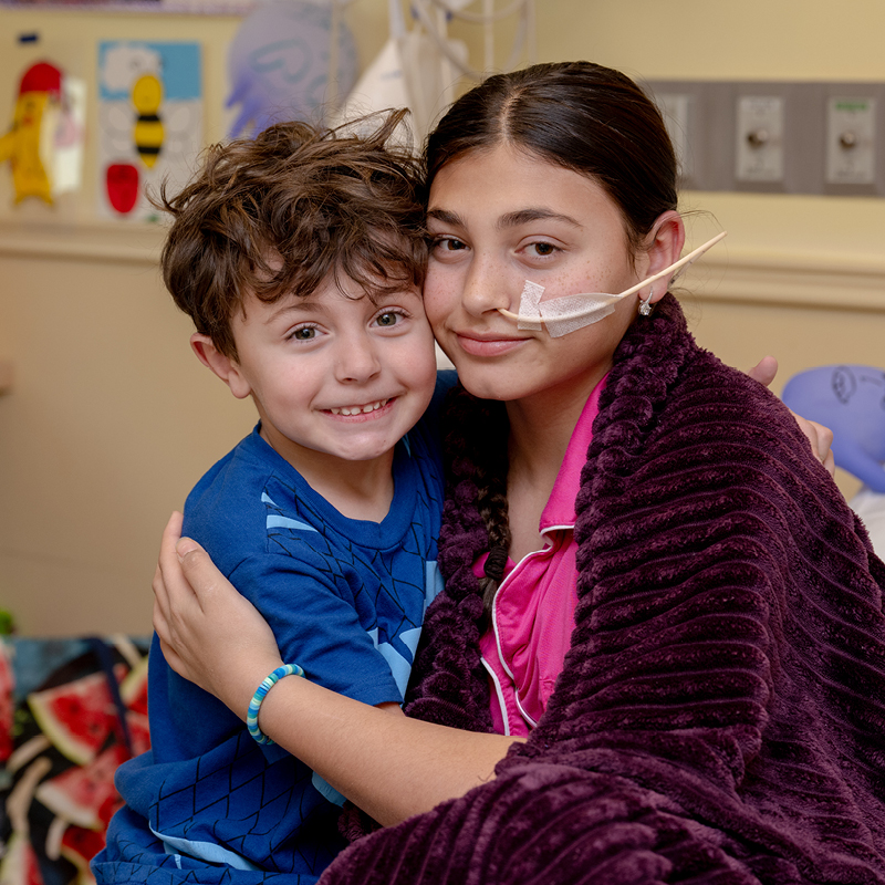 Two children hugging in a hospital room, one wearing a nasal cannula.
