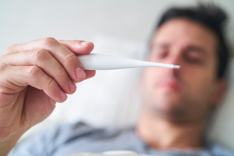 Close up shot of a man holding out a thermometer, taking his temperature while in bed. 