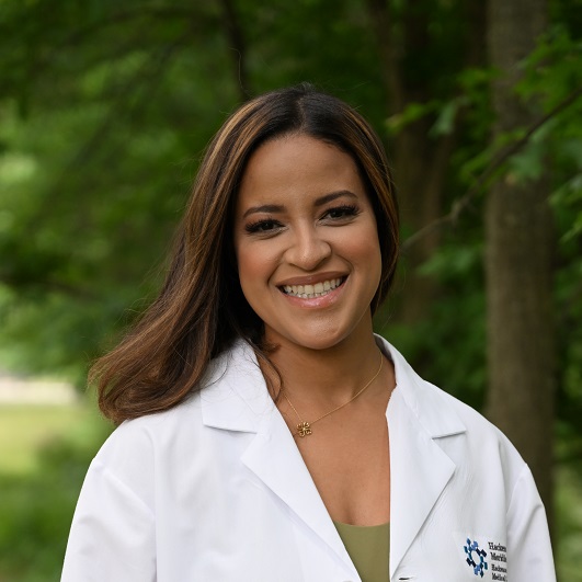 Dr. Magna Pastrano standing out in the park with, with her lab coat on, smiling, in front of trees.