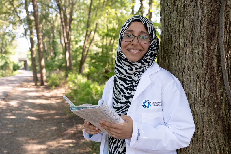 Dr. Iqbal standing outside, next to a tree, holding a pen and journal, smiling.