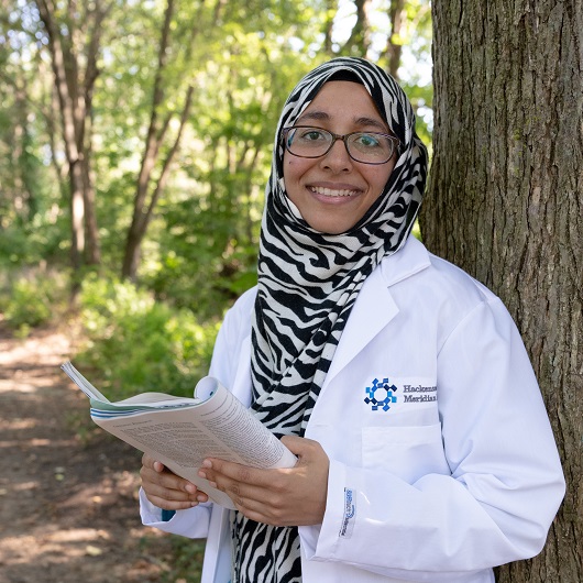 Dr. Iqbal standing outside, next to a tree, holding a pen and journal, smiling.
