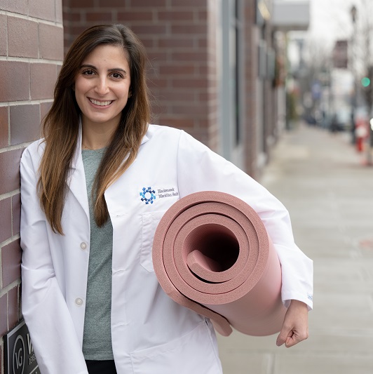 Dr. Sarah Mateen smiling and holding a rolled-up yoga mat.