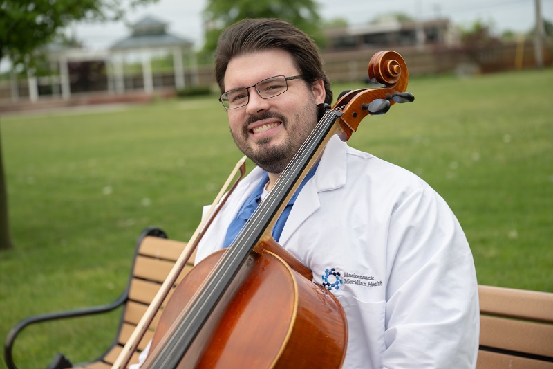 Dr. John Mosko sitting on a park bench smiling, holding a cello. 