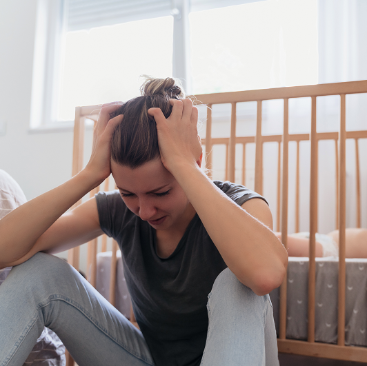 Woman sitting next to her child's crib with her head in her hands - feeling anxious and depressed in postpartum.