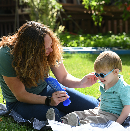 Mother putting sunscreen on her young son while sitting out in the grass on a warm sunny day.