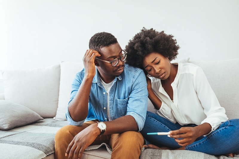 A sad young woman holds a pregnancy test in her hand, sitting on the couch with her partner.