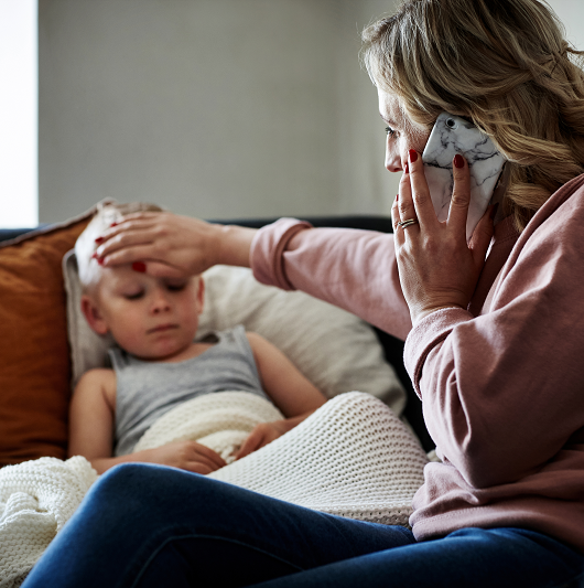 Mother touches her son's forehead as he rests on the couch, feeling for a fever. Young boy not feeling well, possibly sick with RSV.
