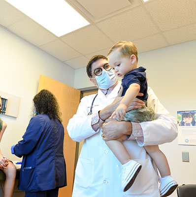 Pediatrician holding a child patient with their older sibling also sitting in the patient room. 
