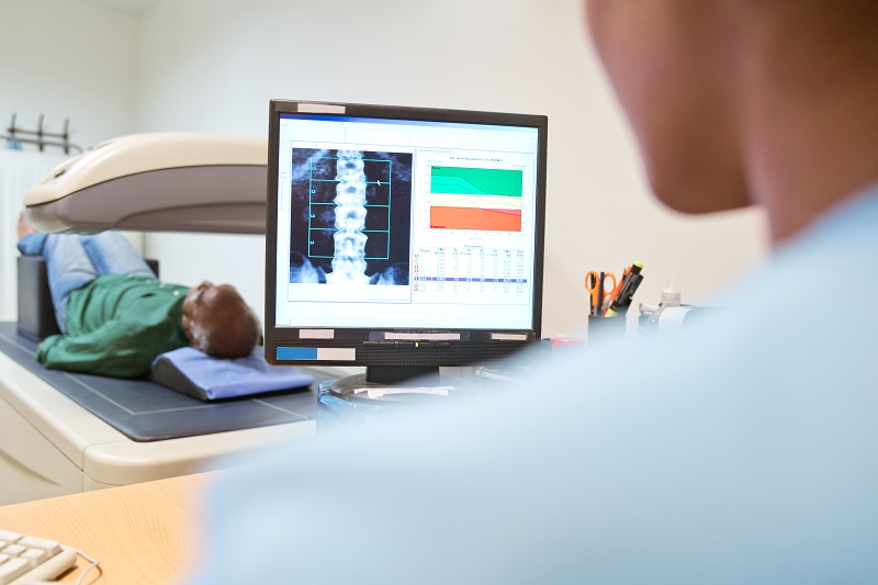 Healthcare worker analyzing x-ray image on computer monitor. Senior male patient is lying on examination table. 
