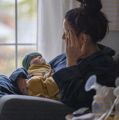 A new, postpartum mother sitting on a rocking chair in her living room wearing a housecoat holding her newborn baby. There is a breast pump and nursing supplies around.
