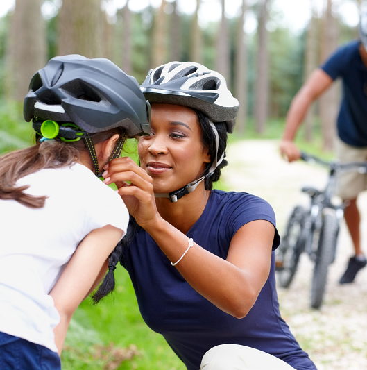 Caring young mother helping daughter put on helmet with father in background, wearing a helmet - Outdoors in country side
