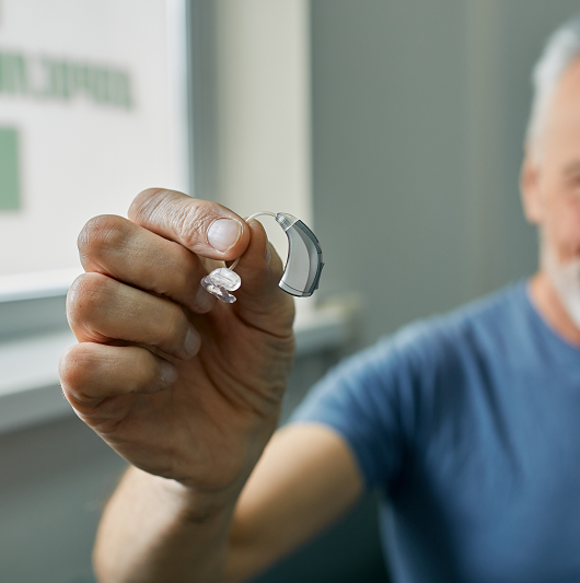 Senior man holding over-the-counter hearing aid in hand on foreground, close-up. Treatment of deafness in elderly people