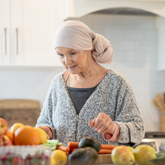 A senior woman with cancer stands at her kitchen counter as she works to prepare an assortment of fruits and vegetables. She is dressed casually in a sweater and wearing a headscarf as she looks over the produce and makes an effort to maintain a healthy diet during her Chemotherapy