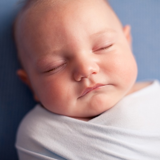 Close up of a sleeping baby lying on his back, swaddled, on a blue sheet.