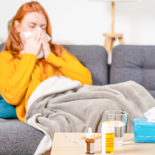 Woman home sick with the flu, blowing her nose on the couch. Over-the-counter pain relievers and medicine on the coffee table.