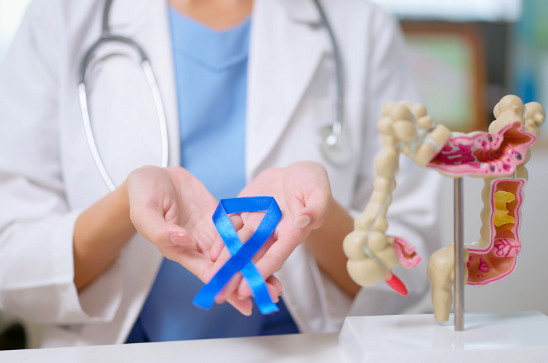 close up of female doctor hand wear white coat holds blue ribbon in front of her chest with colon model on table