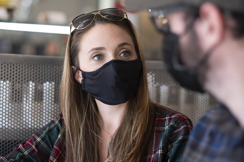 Close up shot of a woman wearing a mask, in a coffee shop, talking with someone. 