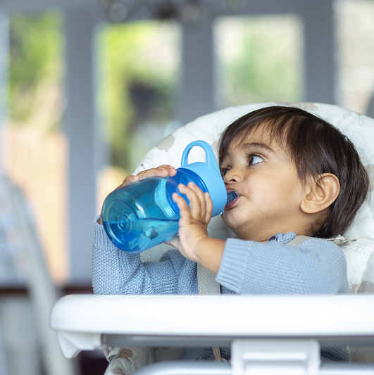 A front-view shot of a young boy drinking water from a reusable water bottle at home, he is wearing casual clothing and sitting in a high chair.