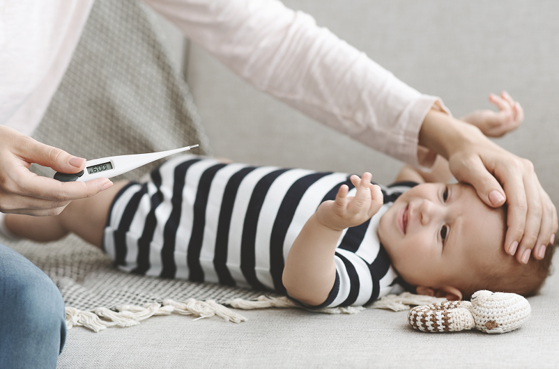 A mother's hand touching her babies forward to feel if it it is warm, while holding a thermometer reading the baby's temperature with a fever of 100 degrees