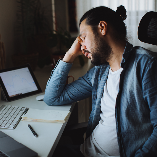 Young man closing his eyes, resting his head on his hand while sitting at his desk in front of his computer at home, struggling to stay awake. Suffering from sleep deprivation. 