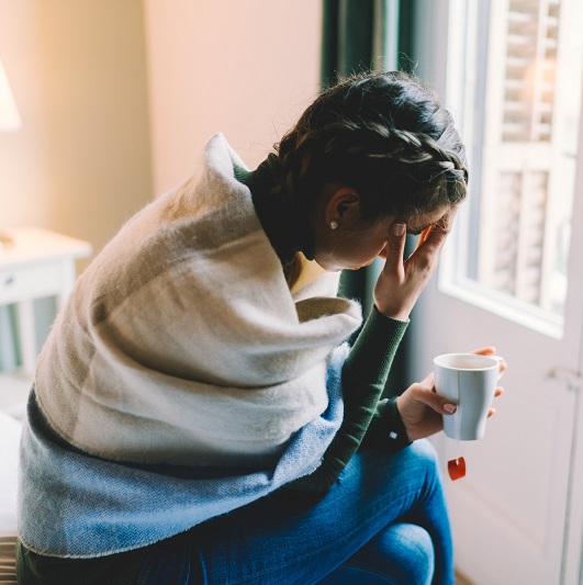 Young woman, sitting on the edge of the bed, bent over with her hand on her face, struggling with depressive symptoms from premenstrual dysphoric disorder.