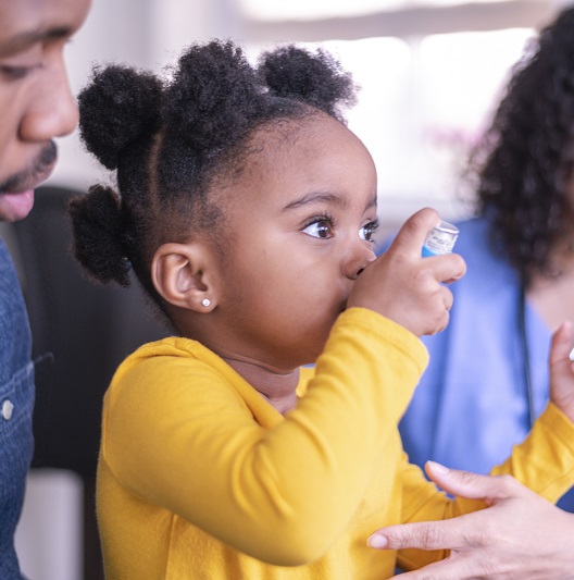 A little girl with asthma is at a doctor's appointment. She is sitting on her father's lap. The girl is using a puffer. The doctor is teaching the child how to use the medical device.