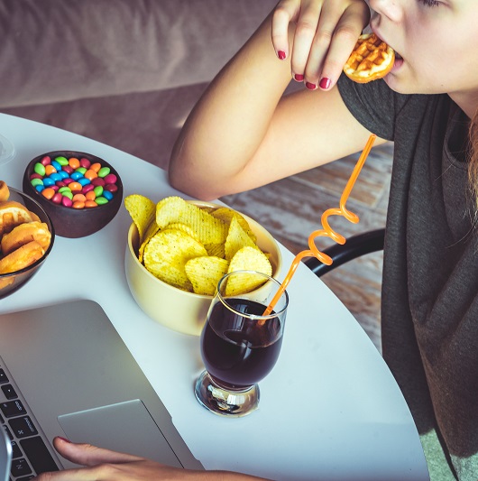 Girl works at a computer and stress eats. Unhealthy food: chips, crackers, candy, waffles, soda.