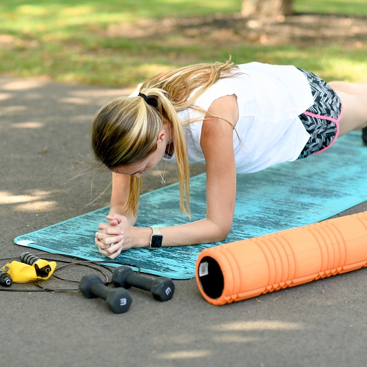 Woman doing a plank on a yoga mat outside in the park, with weights and a jump rope nearby.