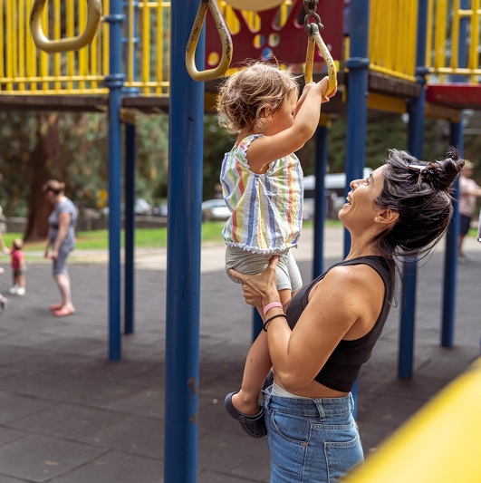 Mom helps a two year old girl use the jungle gym at the playground on a warm sunny afternoon.