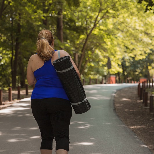 Woman walking away from the park and carrying exercising mat on her shoulders