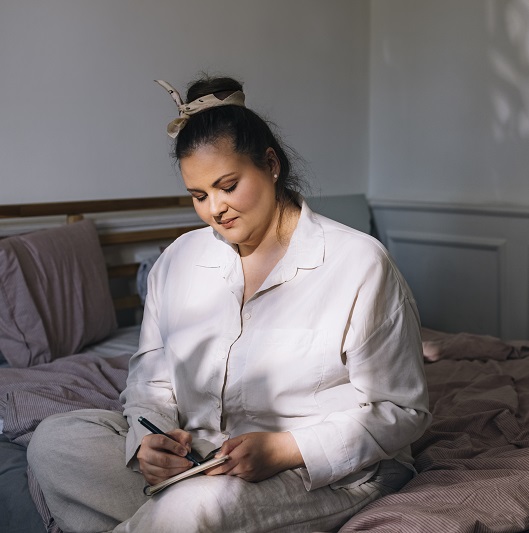 Woman sitting on her bed, in her pajamas taking note of her sleep.