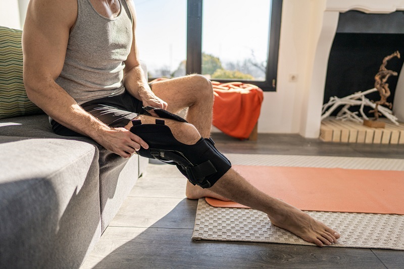 Young man sitting on a sofa putting on a knee brace for an ACL injury.
