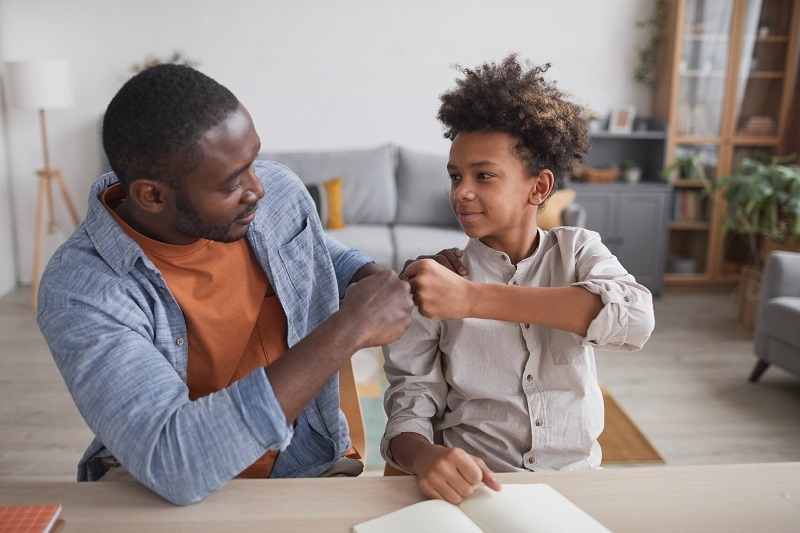 A father and school-aged son fist-bumping and smiling in their home.