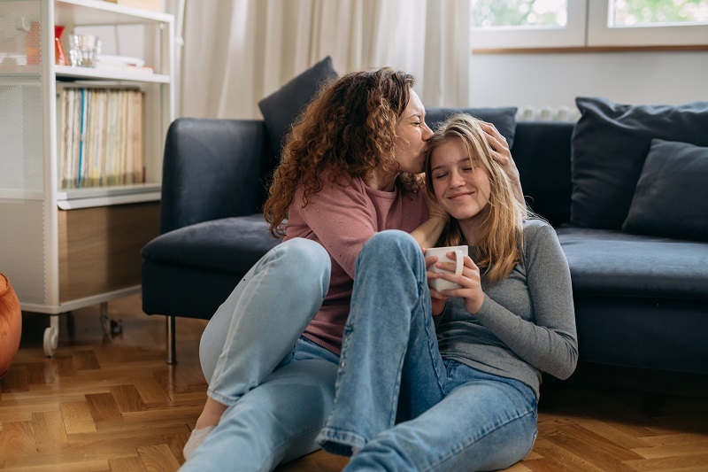 Mom and preteen daughter are sitting next to the couch in their living room, mom is giving her daughter a kiss on head while daughter smiles.