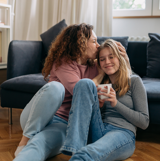 Mom and preteen daughter are sitting next to the couch in their living room, mom is giving her daughter a kiss on head while daughter smiles.