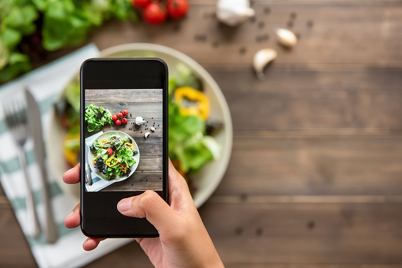 Overhead view of a hand holding a cell phone, taking a picture of a mixed fresh green salad. 