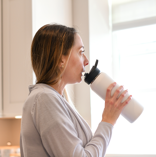 Woman drinking from a stainless steel water bottle, looking out the window. 