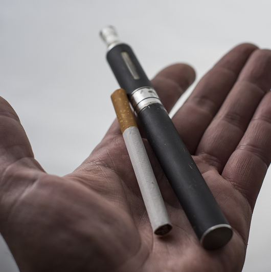 A man's hand holding a vaping cigarette and a traditional cigarette.