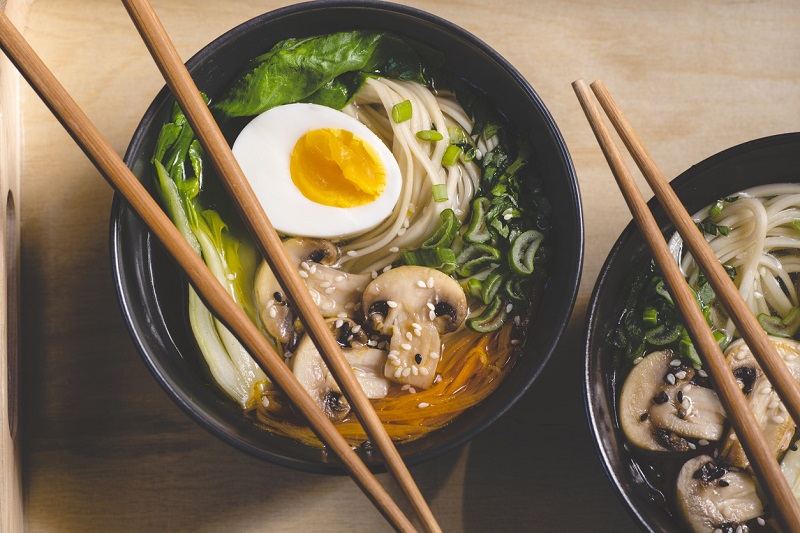 Ramen soup with mushrooms, bok choy, and greens in two black bowls on an orange background.