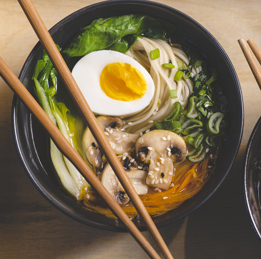 Ramen soup with mushrooms, bok choy, and greens in two black bowls on an orange background.