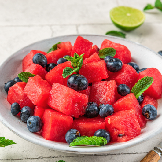A white bowl is filled with fresh watermelon chunks and blueberries that are sprinkled with mint leaves.