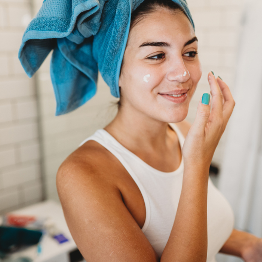 A young woman with her hair in a towel dabs a skincare product on her face.
