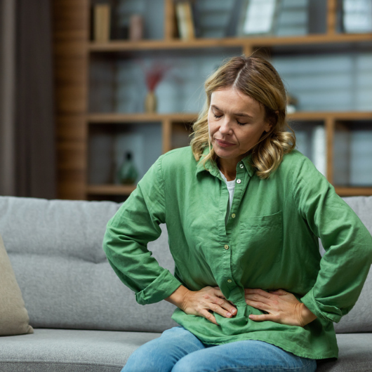 A woman is sitting on the sofa at home, holding her stomach with her hands.