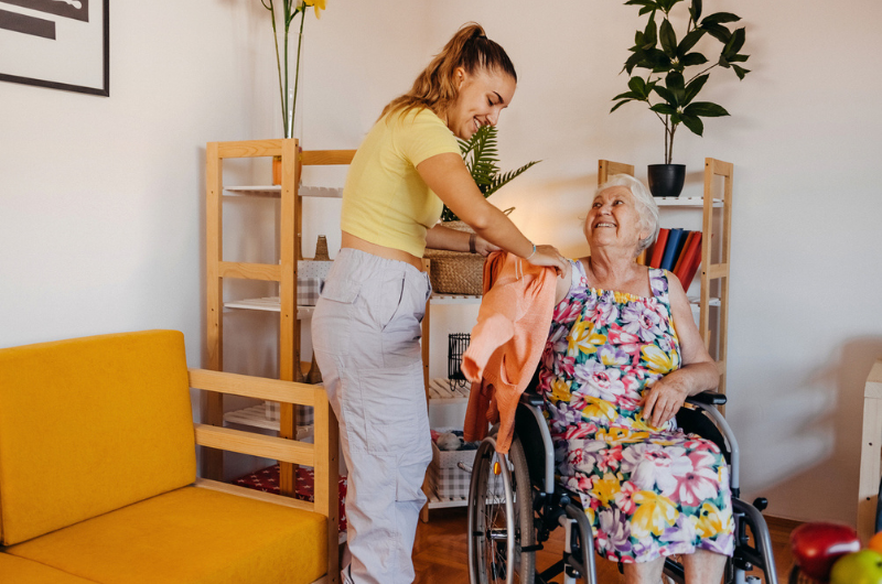 A young woman helps an older woman in a wheelchair put on her sweater.
