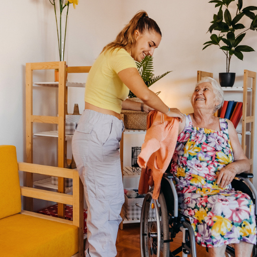A young woman helps an older woman in a wheelchair put on her sweater.