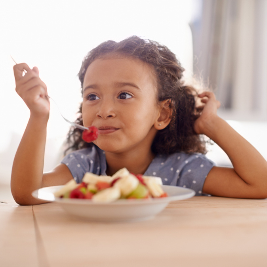 Brunette little girl eating fruit salad at a table.