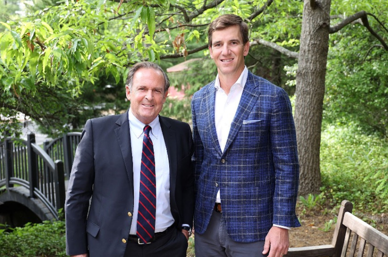 Peter Cancro and Eli Manning stand side-by-side and smile at the camera with greenery behind them.