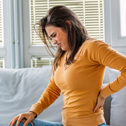 Portrait of a young brunette woman sitting on the couch at home with back pain.